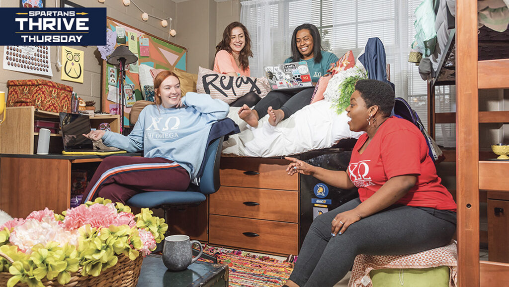 Four UNCG students sit on chairs or on a bed while talking in a dorm room.