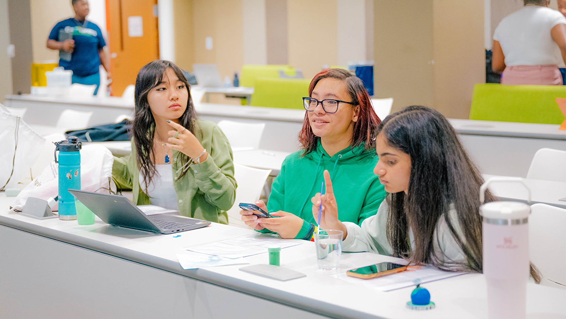 Three high school students sit together at a long table with their laptops and take notes.