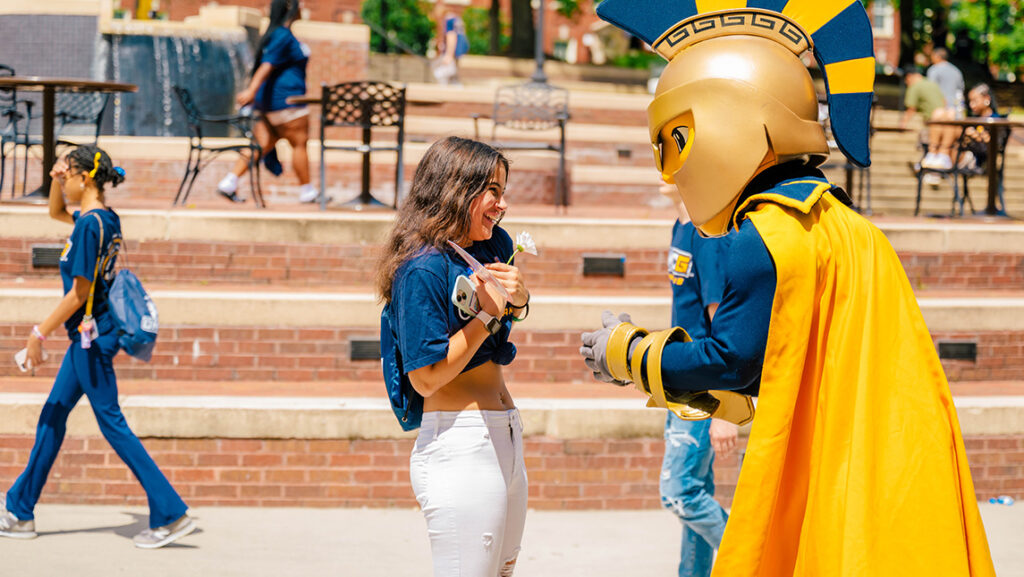 A new student to UNCG holding a daisy talks with Spiro the mascot.