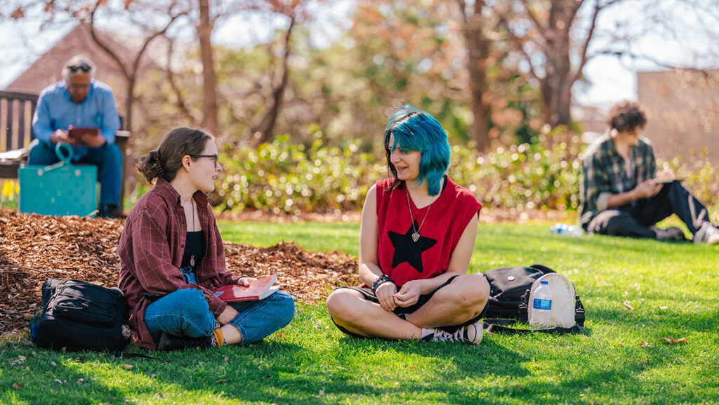 Students sit on a green lawn on a UNCG campus quad.