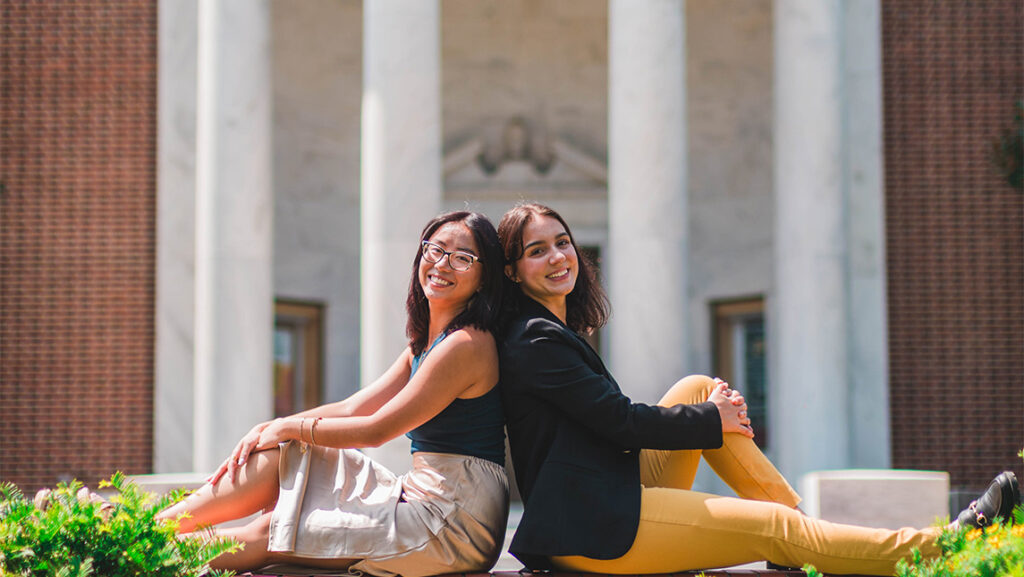 Two students sit back-to-back outside of the library.