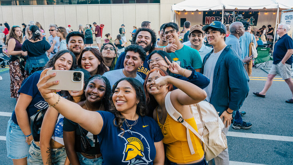 Group of UNCG students pose for a selfie in a downtown street withe people walking in the background and Folk Fest tent behind them.