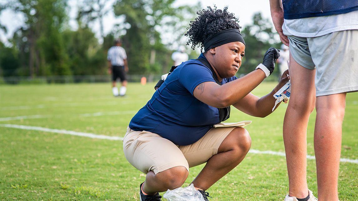 An athletic trainer helps tend to a person's leg with a field behind them.