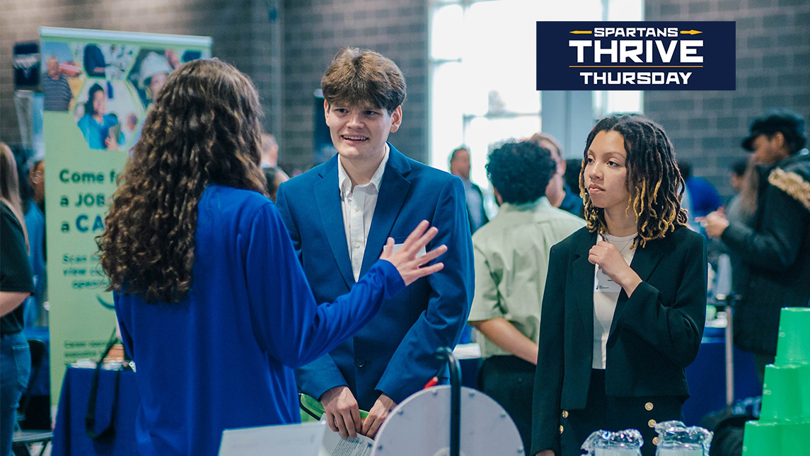 Students dressed in business suits talk to an employer at a career fair at UNCG. Spartans Thrive Thursday logo is in the top right corner.