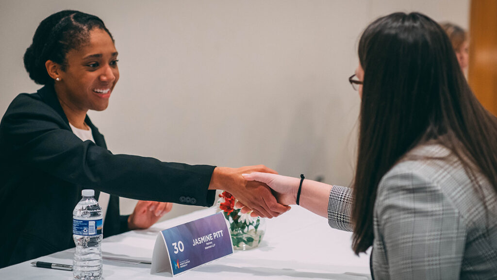 Two women dressed in business suits shake hands across a table they are sitting at.