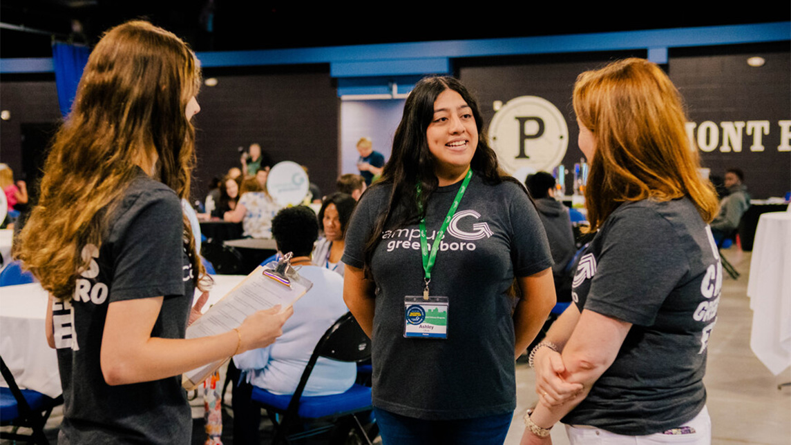 Three students wearing matching t-shirts talk at an event.