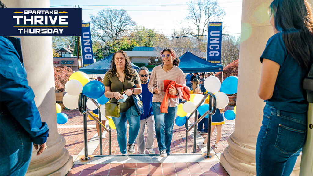 A mother and daughter walk up the steps to a door held open by UNCG volunteers.