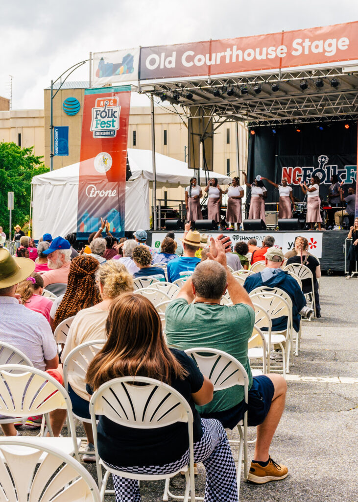 Stage set up in a Greensboro downtown parking lot with a group of singers on stage and a crowd watching from folding chairs. 