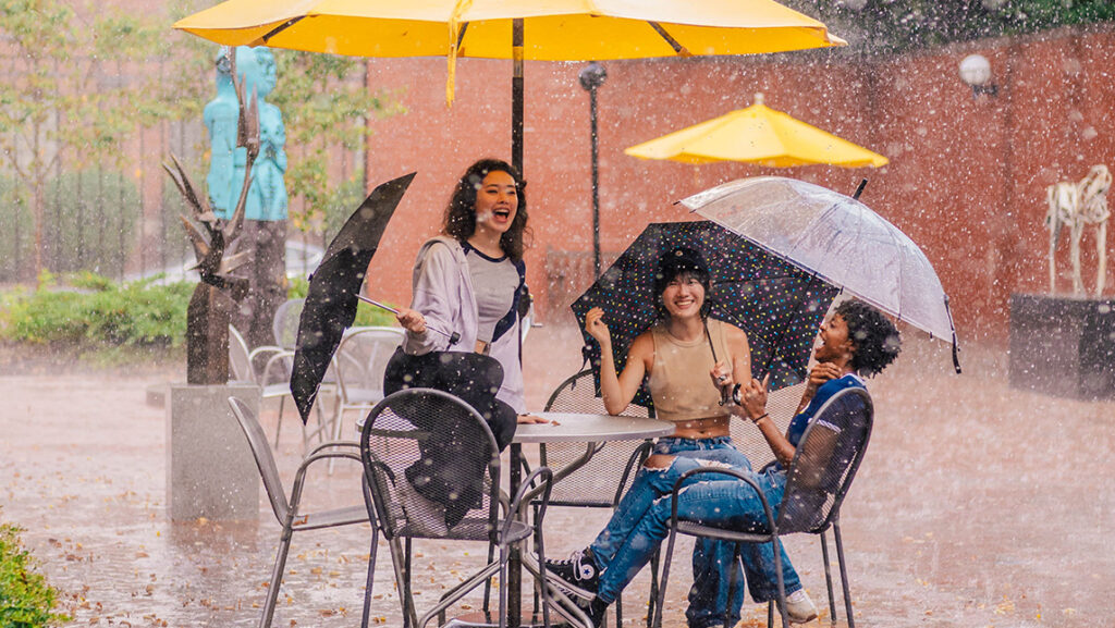 A group of students laugh in the rain as they sit in the UNCG Weatherspoon courtyard.