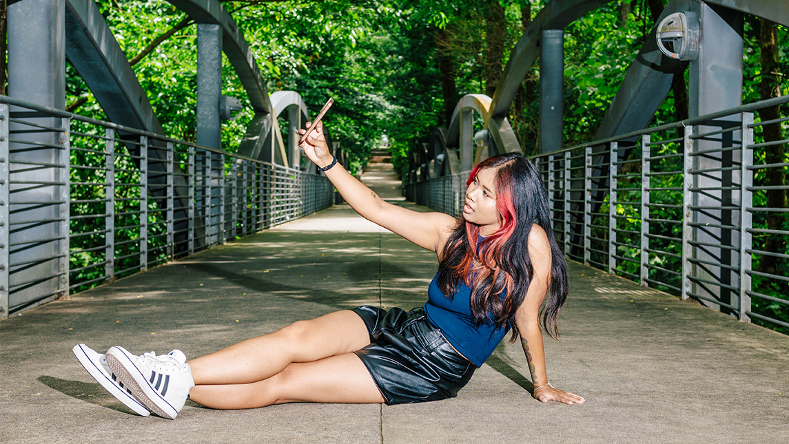 A student sits on the walkway of the UNCG bridge and takes a selfie.