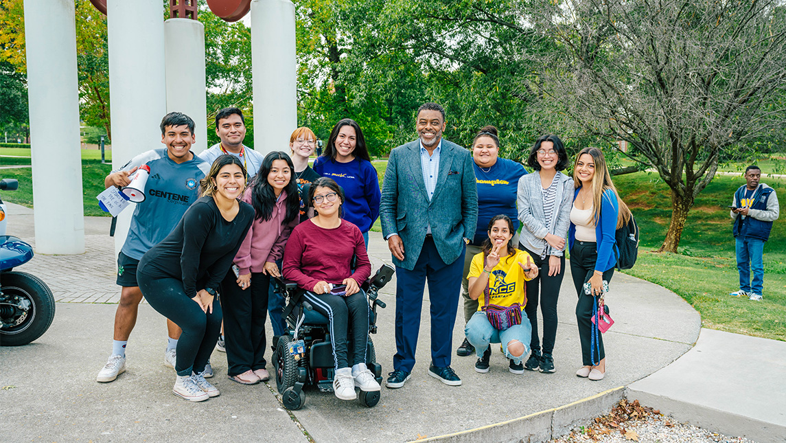 A group of UNCG students stand with Chancellor Gilliam beside the clock tower.