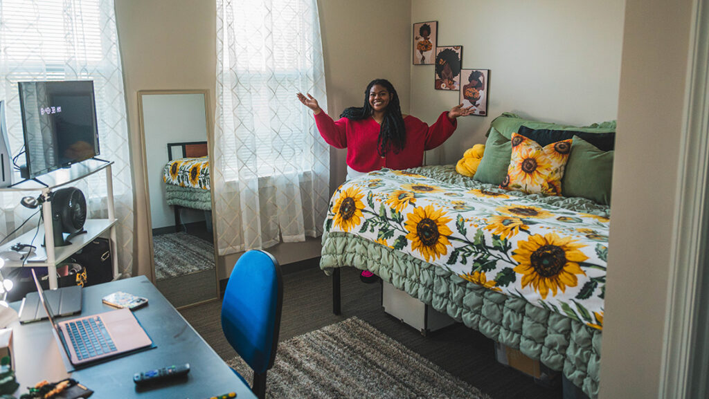 Girl poses in her dorm room decorated with sunflowers and holds her arms out. 