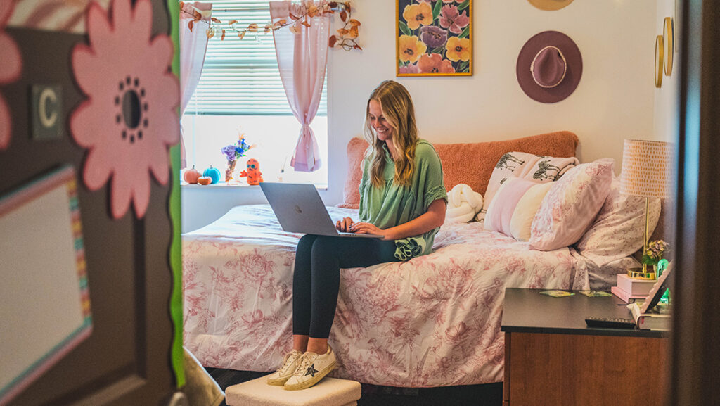 Student sits on her bed working on a laptop in a room decorated in flowers and cowboy hats. 