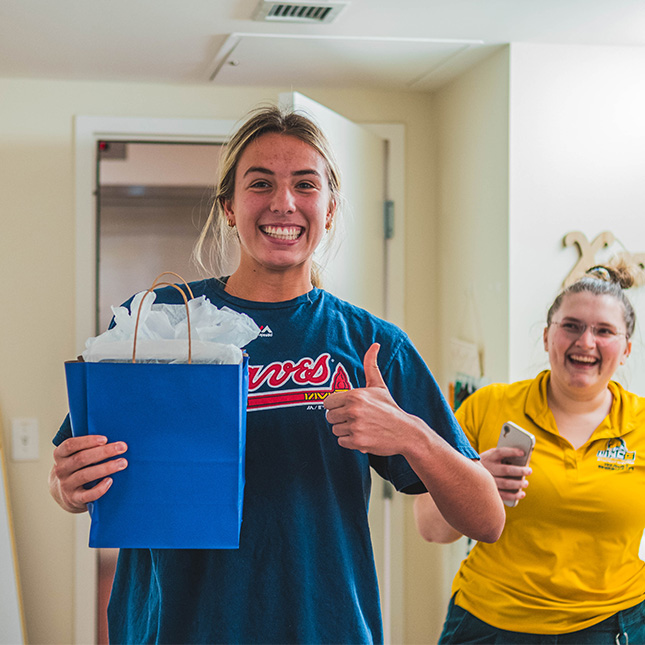 Girl holds a gift bag and gives a thumbs up while a woman in a UNCG shirt smiles at her from behind. 