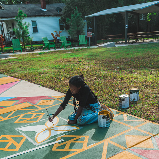 Student paints a sidewalk mural in a park.