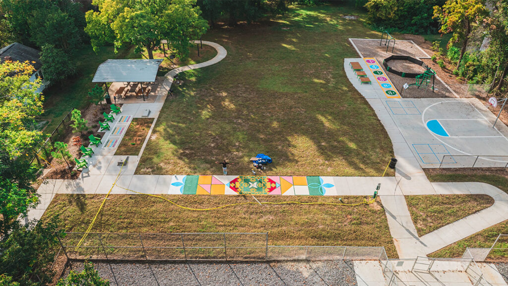 Wide arial shot of a park with trees around it, lawns separated by sidewalks, a picnic area, playground and basketball court. In the center a section of the sidewalk is painted colorfully and you can barely see the artist standing beside it with her arms outstretched.