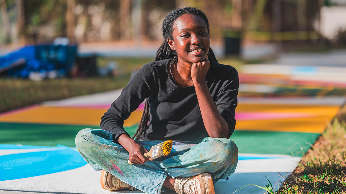 Girl sits cross-legged on a painted park sidewalk with a paint brush.