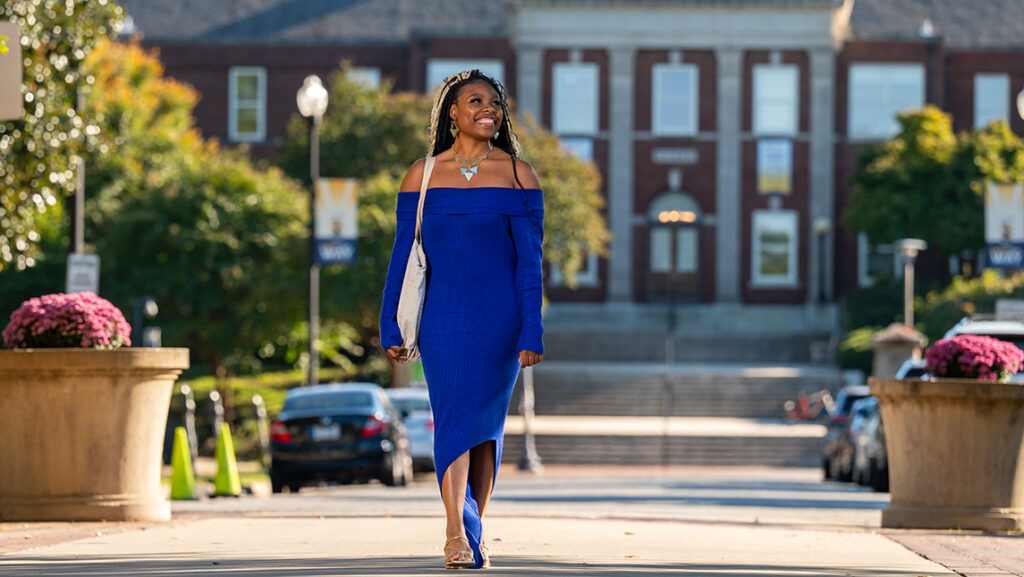 Student in blue dress walks down College Ave at UNCG with the Curry building behind her.