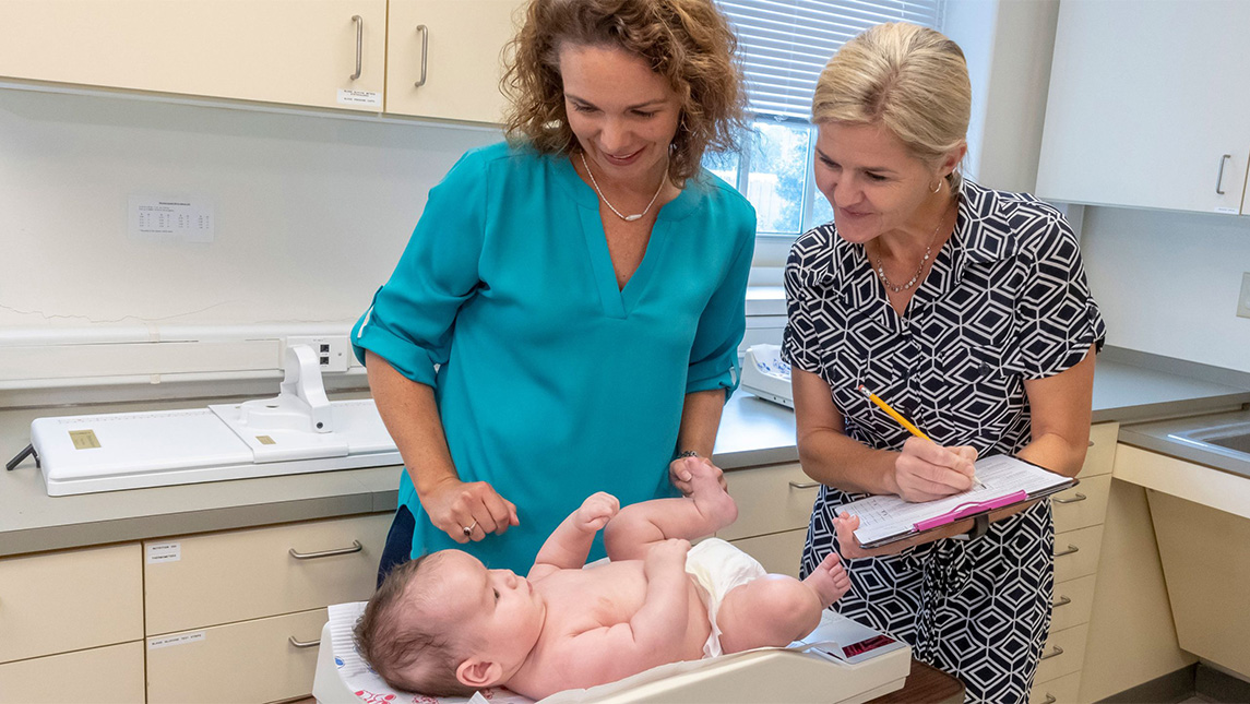 Two researchers smile at an infant while collecting data with a medical setting in the background.