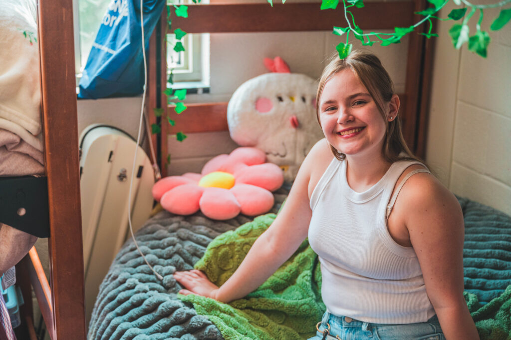 Student sits on a bed with soft pillows and ivy greenery decorating the walls.