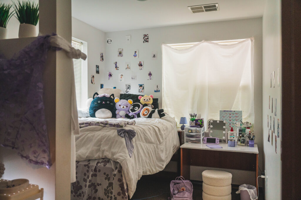 Dorm room decorated with photos on the walls, plushies on the bed, and a dressing table.