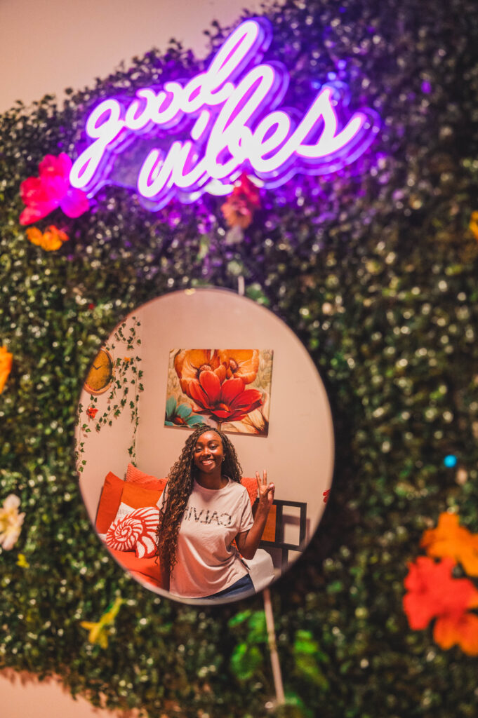 Student sits on her bed with orange bedding and orange flowers in artwork as seen in a mirror hanging on the opposite wall with greenery and a "good vibes" purple neon sign over it. 