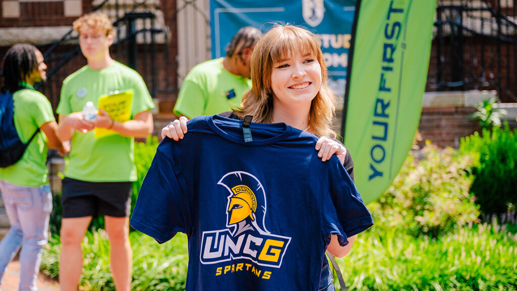 Student stands in front of Forney building posing with a UNCG t-shirt held in front of her. Your First Year banners and students in lime green shirts are behind her.