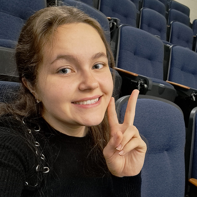 Student gives a peace sign in a selfie taken in an auditorium.