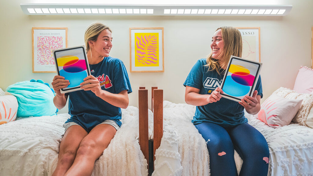 Girls sit on beds that are positioned end to end on a dorm wall and hold ipads.