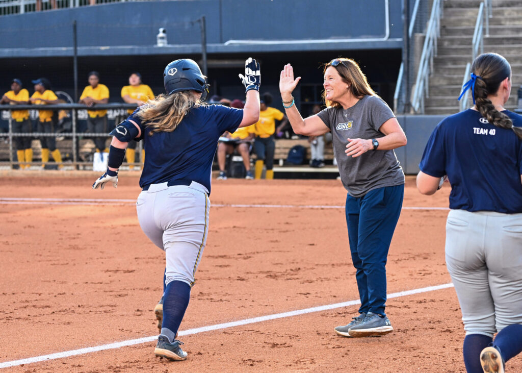 Softball player high fives a coach as she runs bases with the dugout full of players in the background.