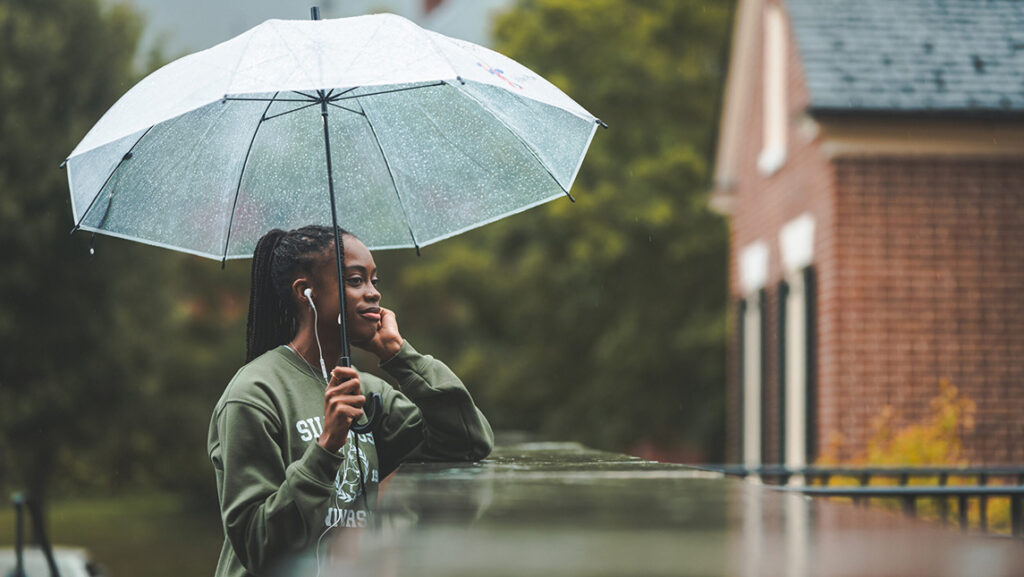 A young student stands in the rain with an unbrella as she leans on a wet ledge and looks across the campus.