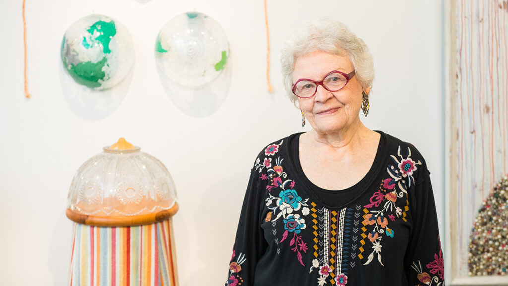 Artist Carol Cole Levin stands beside art exhibits.