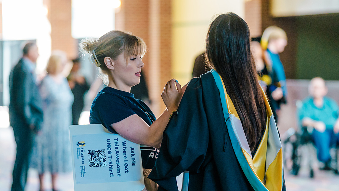 A UNCG volunteer helps a graduating student with her Commencement robe.