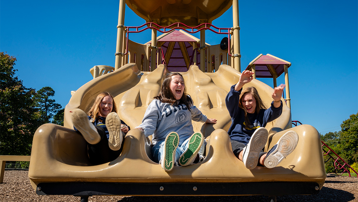 UNCG students Brooke Parks, Mary Grace Smith, and Sidney Cassidy laugh as they slide down a playground slide.