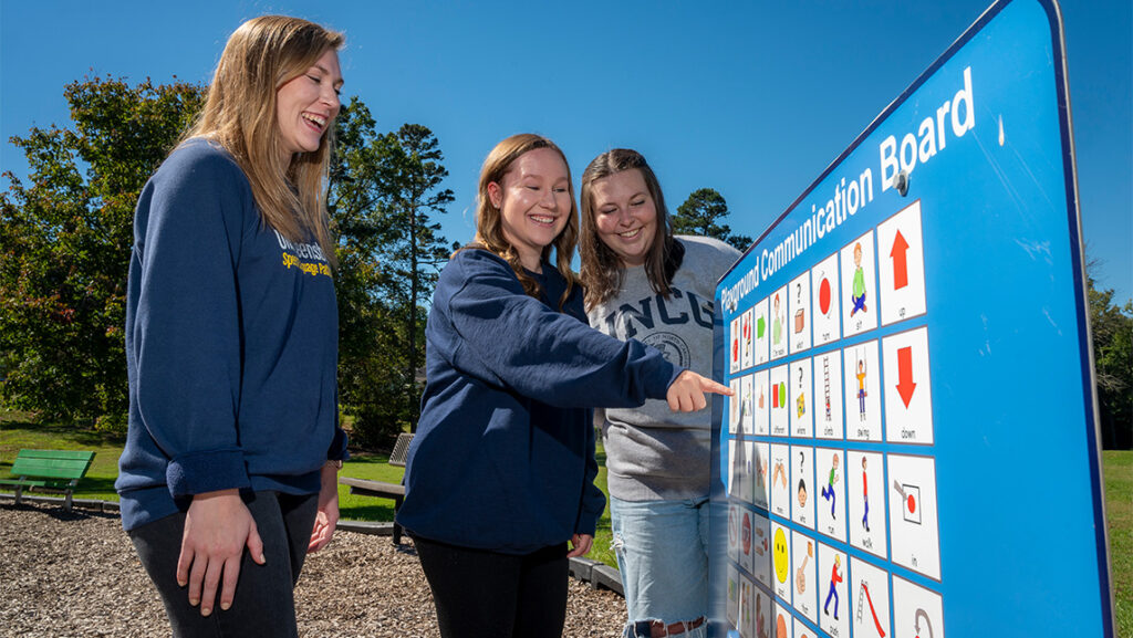 Three UNCG students point at pictures on a board at a playground.