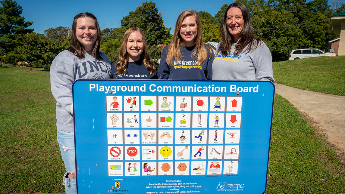 UNCG students Mary Grace Smith, Brooke Parks, and Sidney Cassidy stand behind a picture board with their professor Emily Hamuka.