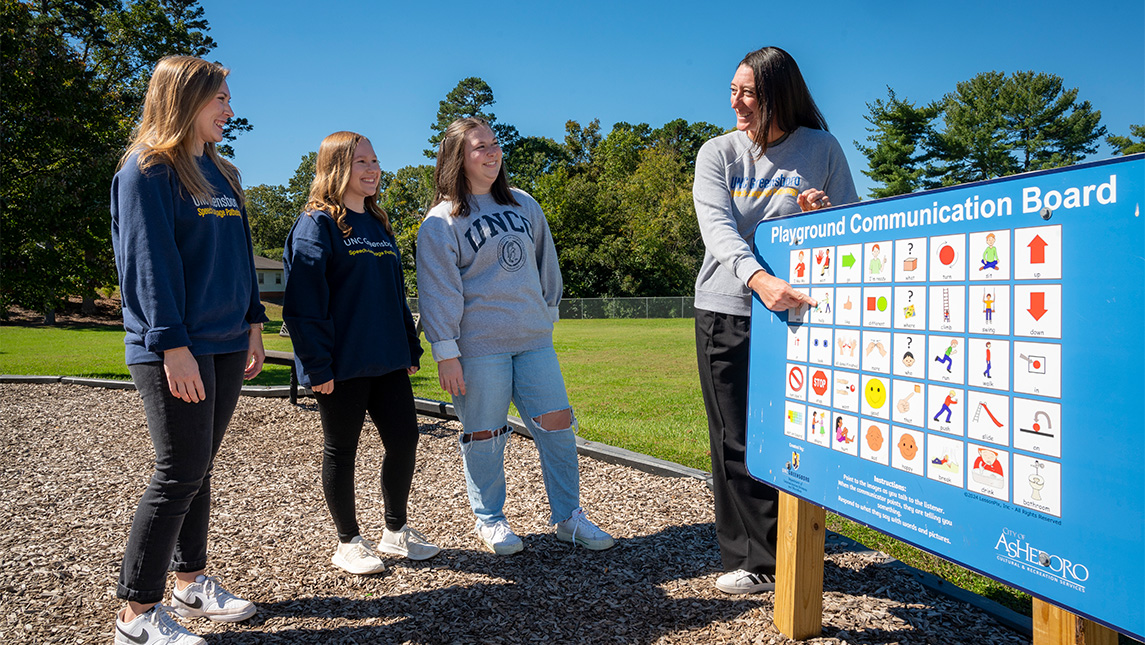 UNCG Dr. Emily Hamuka points at a picture board in a park to her three students Sidney Cassidy, Brooke Parks, and Mary Grace Smith.