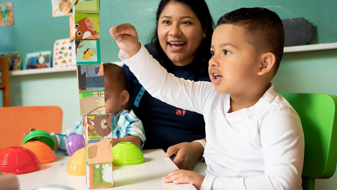 A little boy builds with blocks on a table while an early childhood education professional cheers him on.