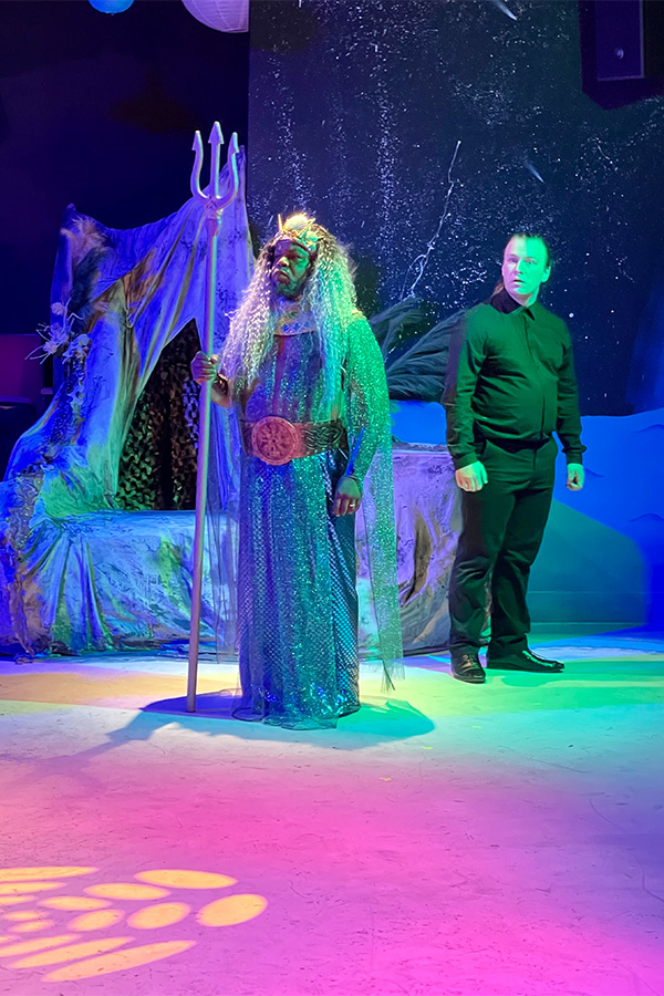 An actor playing King Triton in The Little Mermaid stands onstage beside an interpreter wearing black clothes.