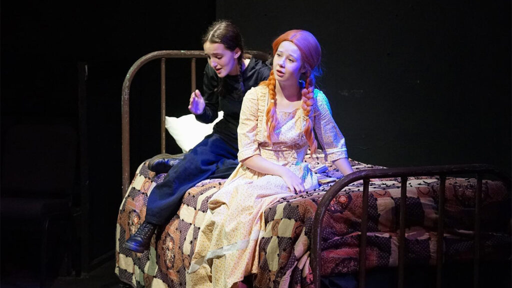 An actress playing Anne of Green Gables sits on a bed while a UNCG student in black clothes shadows her performance.