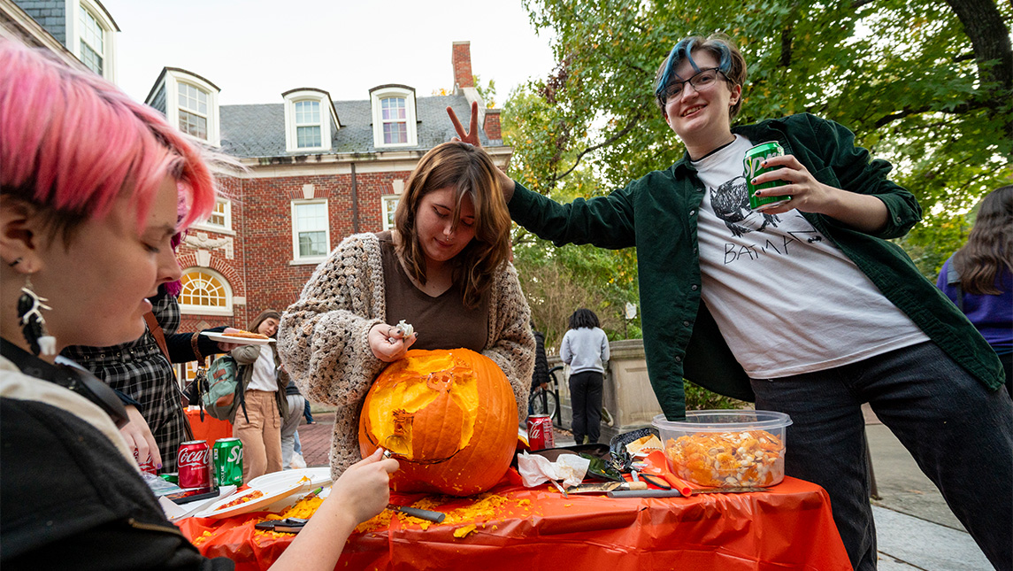 UNCG Residential College student puts bunny ears over another student who is carving a pumpkin.