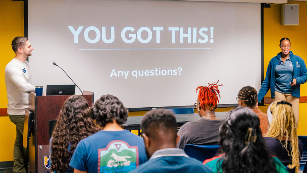 Class of students with two advisors at the head of the class standing at a podium with a screen that reads "You Got This!"