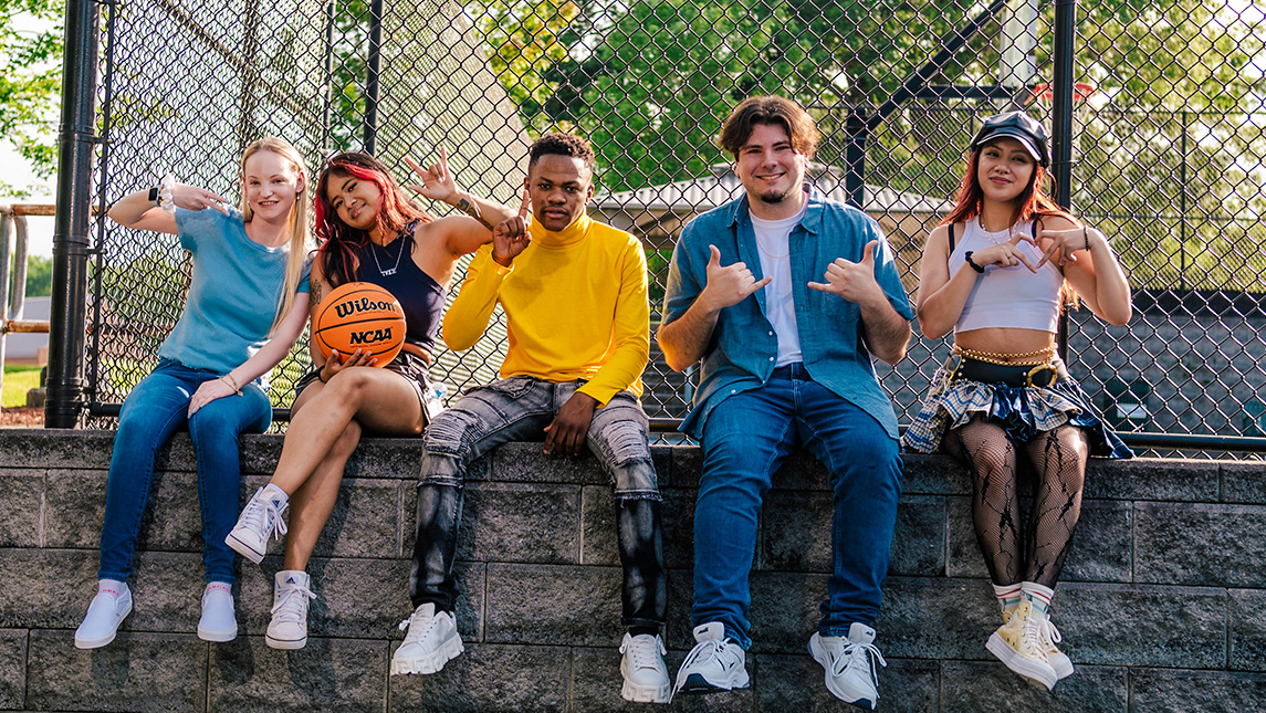 UNCG students sit on a ledge with a fenced basketball court behind them and give hand signals (hearts, thumbs up, number 1).