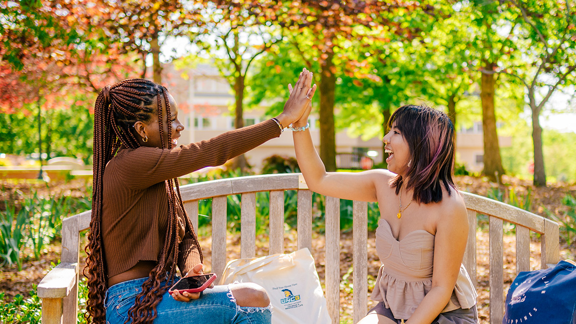 Two students sit on a bench on UNCG's campus and high five each other.