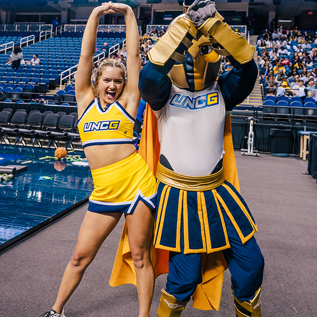 UNC cheerleader bumps hips with Spiro mascot on the sidelines of a basketball game at the Greensboro Coliseum.