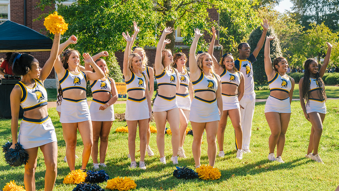 Smiling UNCG cheerleaders stand in a line in a campus quad with hands outstretched and pompoms around them.