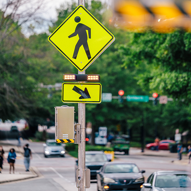 Yellow crosswalk sign with traffic lined on a campus street in the background and students walking on sidewalks.