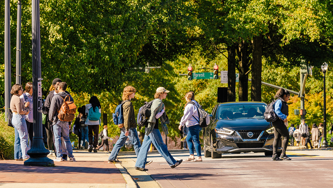 Students crossing a busy intersection at UNCG with trees lining the streets and cars waiting for them to cross.