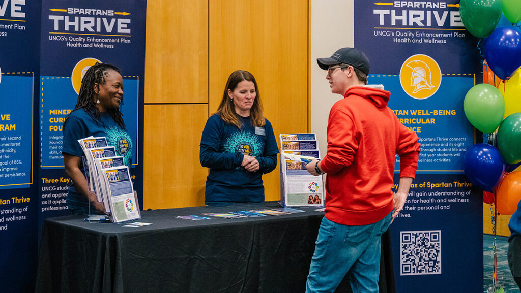 UNCG Student Success staff talk to a student beside a "Spartans Thrive" banner.