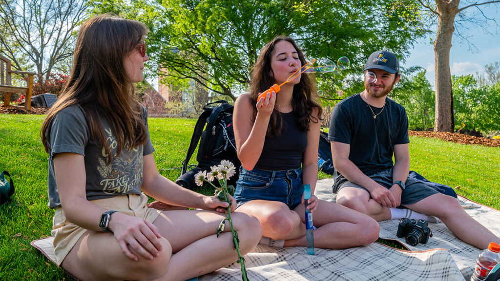 Students blow bubbles while sitting on a blanket of the UNCG lawn.